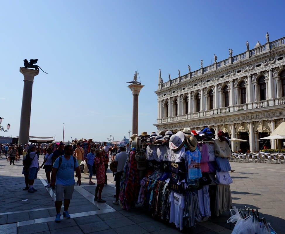 People walk past a souvenir stall in St. Mark's Square in Venice, with the Doge's Palace and two tall columns in view.