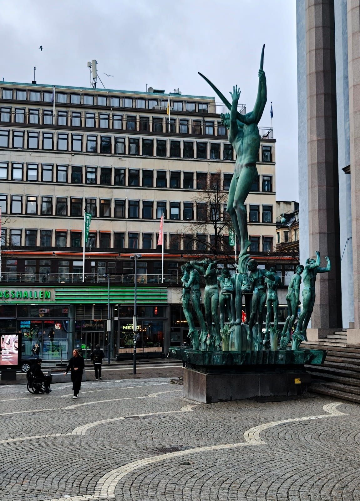 Bronze fountain sculpture in a city square, with a large building and pedestrians in the background