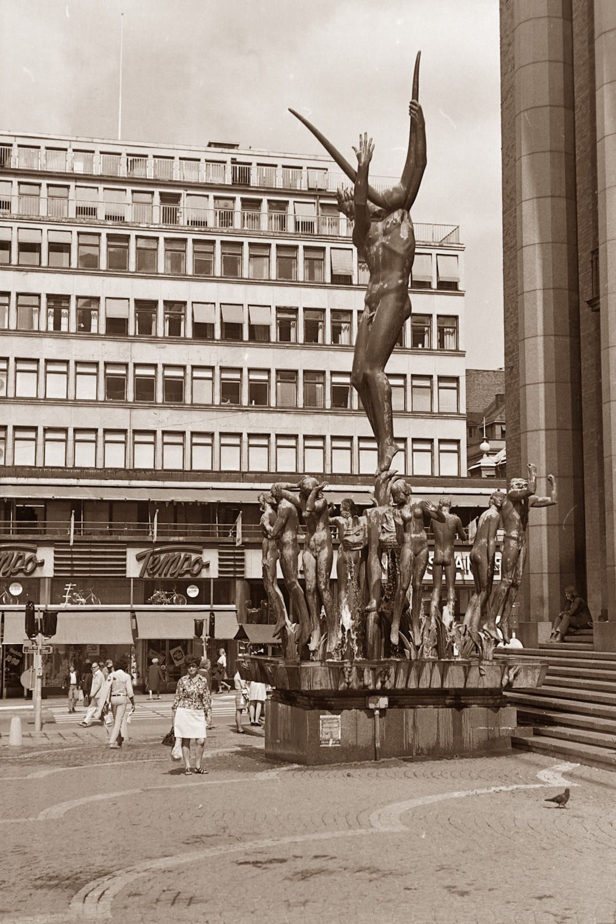 Sepia city square with a bare tree sculpture, a building facade, and a few pedestrians below