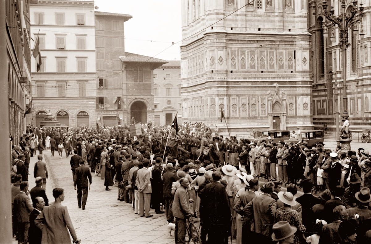 A crowd gathers in a paved square in Florence near Giotto's Campanile, capturing a historic gathering from a low angle.