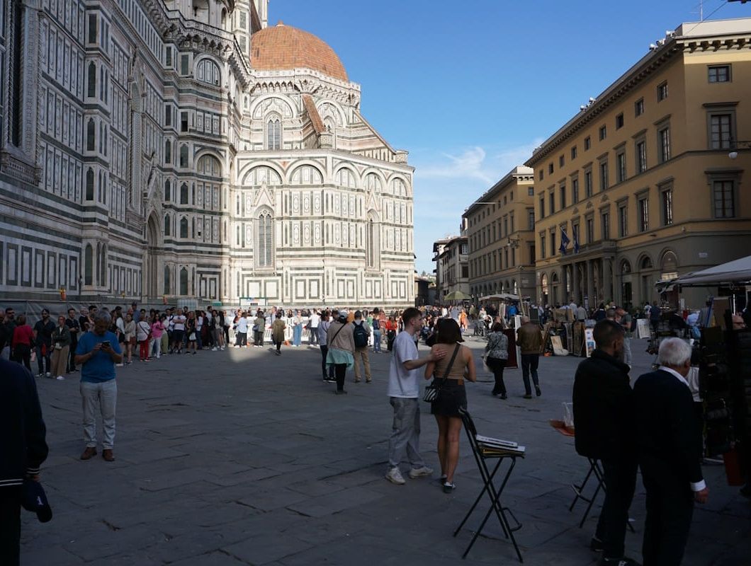 People walk in a sunlit plaza beside the historic, white-and-green marble exterior and tiled dome of the Florence Cathedral.