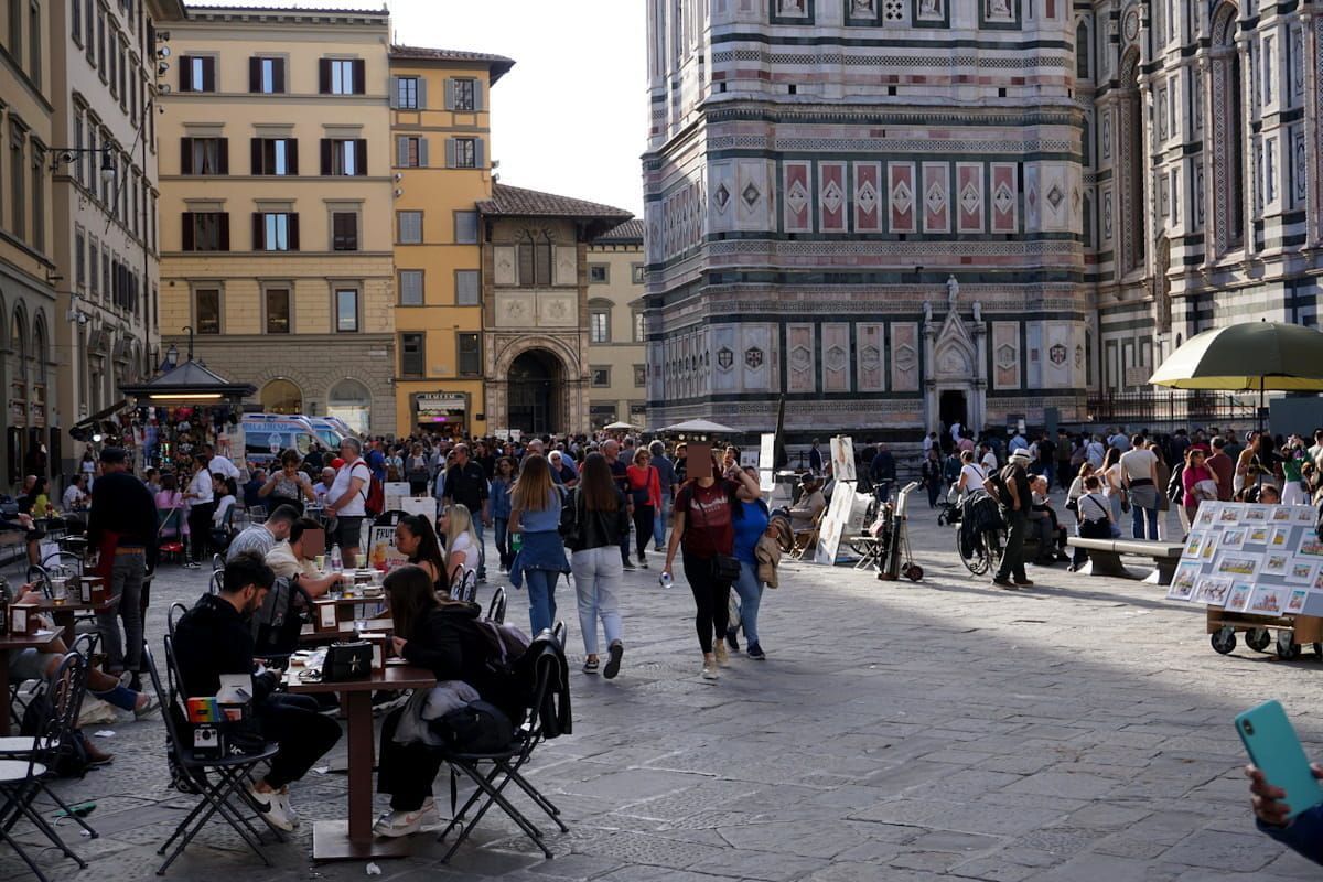 A bustling outdoor square in Florence, Italy, with cafes, pedestrians, and the ornate facade of the Duomo in the background.