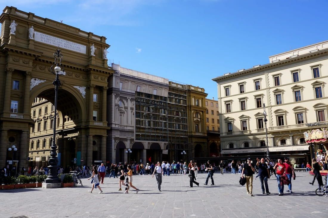 A large, sunlit cobblestone square in Florence, Italy, featuring historic archways and people walking under a blue sky.