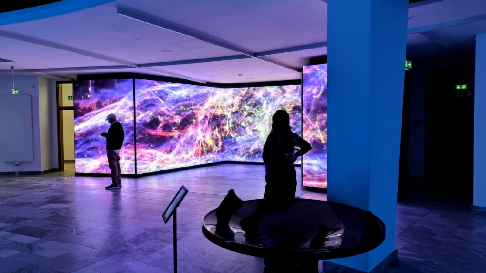 Zeiss-Großplanetarium People viewing a colorful, abstract art installation on large screens in a museum space, lit with blue hues.