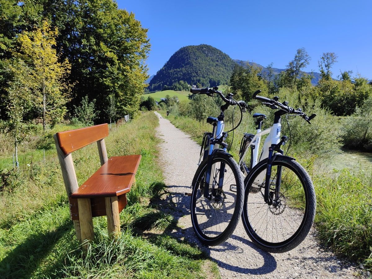 Two bikes parked on a path next to a bench, mountain in the background. Sunny day.