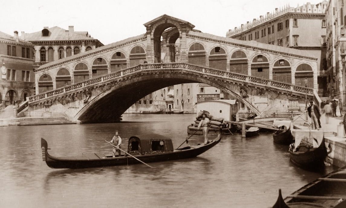 A vintage black-and-white photo of the stone Rialto Bridge over the Grand Canal in Venice, with gondolas in the water.