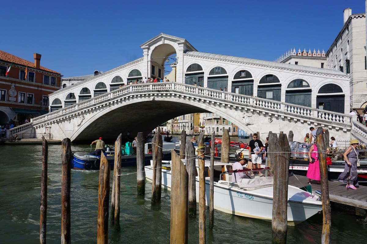 The white stone Rialto Bridge spans a canal in Venice, with wooden mooring poles and boats in the foreground.