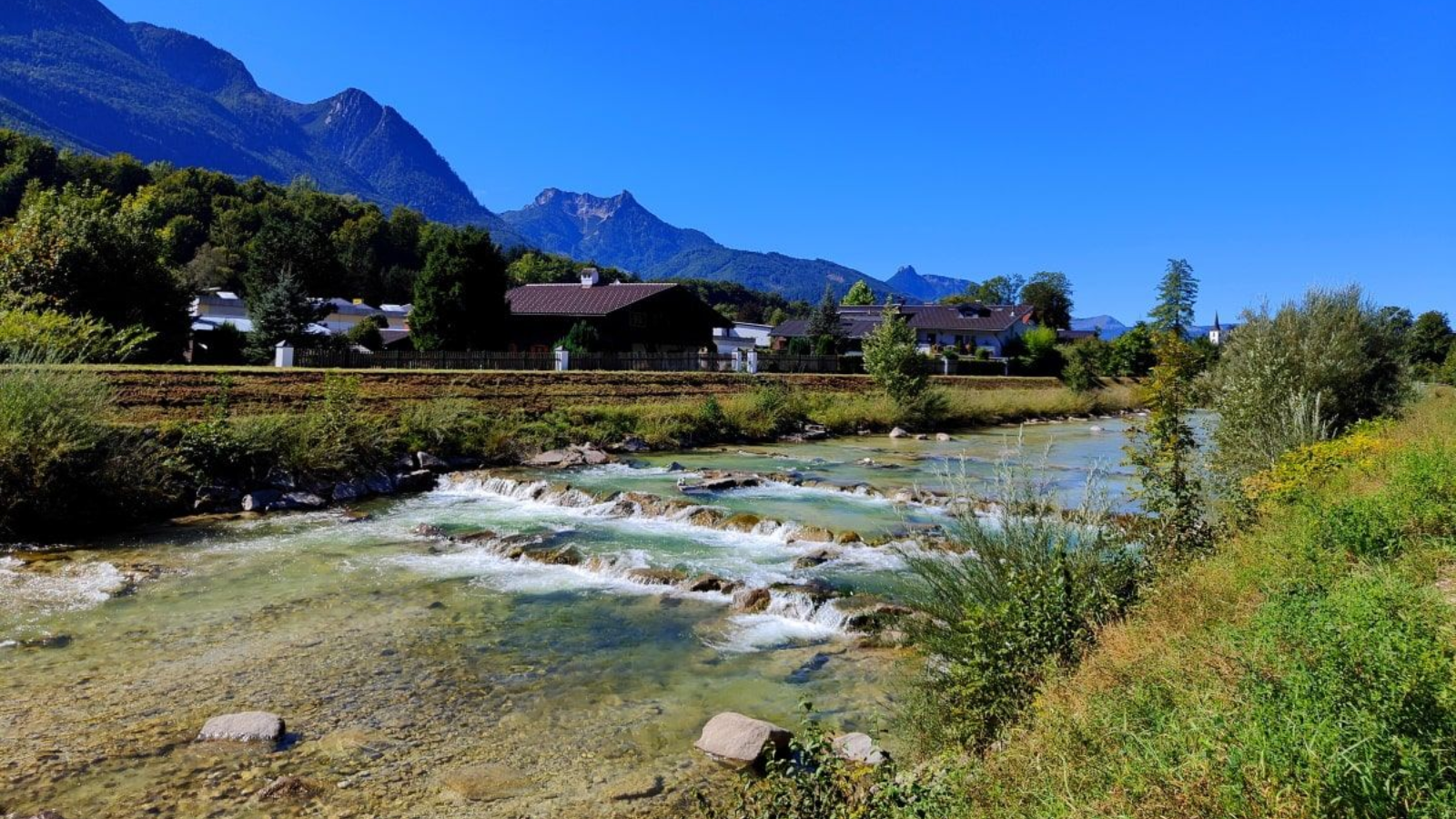 A river flows past a village and mountains under a clear blue sky.