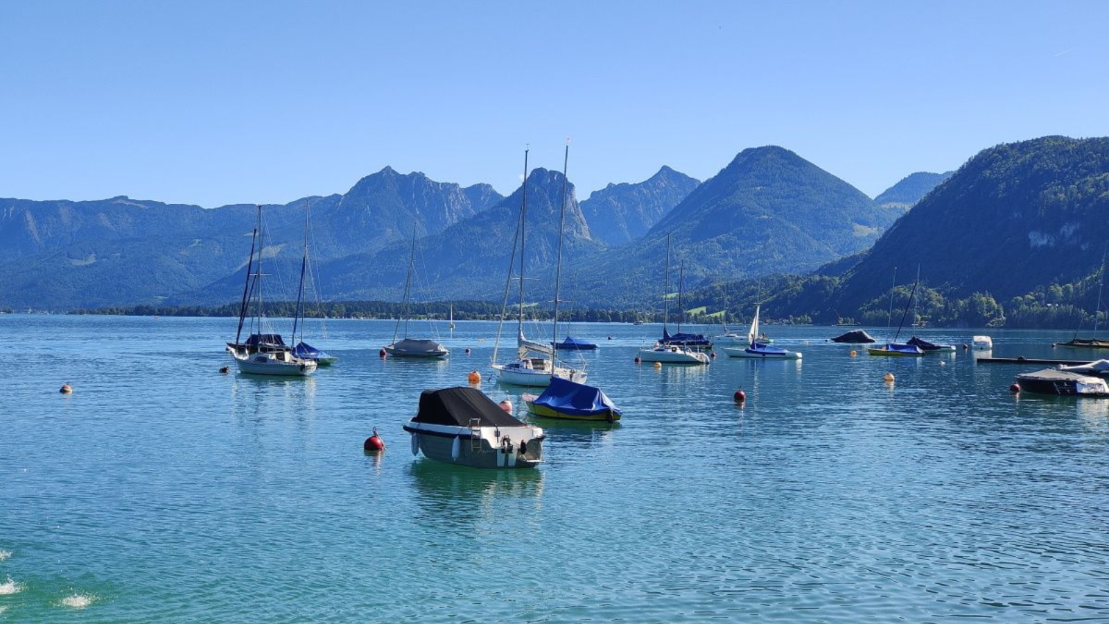 Boats bob on turquoise water in a lake, backed by green mountains under a clear blue sky.