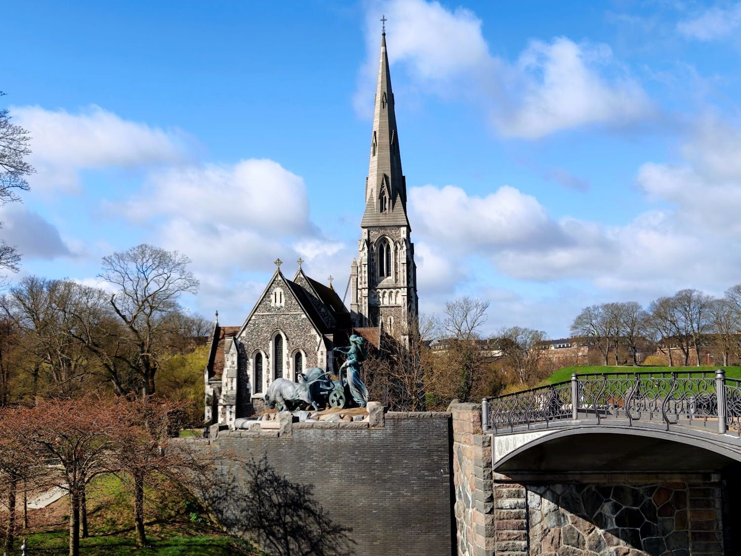 Stone church with tall spire beside a bridge, under a blue sky with clouds and trees.