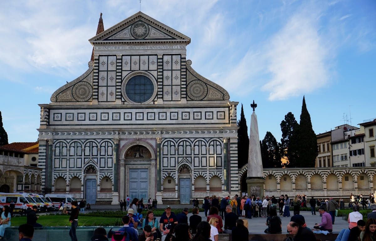 The ornate, green and white marble facade of Santa Maria Novella church in Florence, with a crowd in the plaza below.