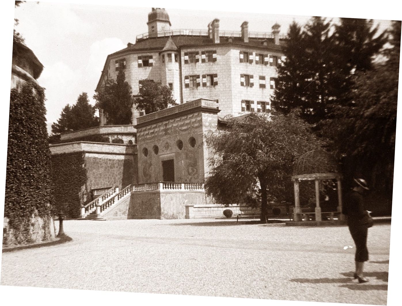 Stone building with a large castle in the background. Stairs, trees, and courtyard in view.