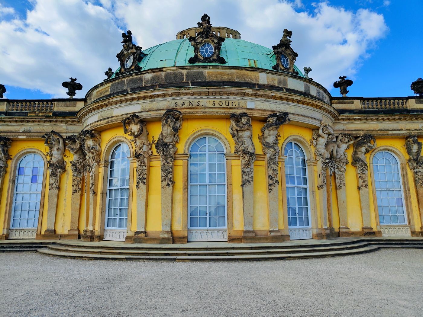 Yellow palace facade with arched windows, blue dome, and steps, set against a blue sky with clouds.