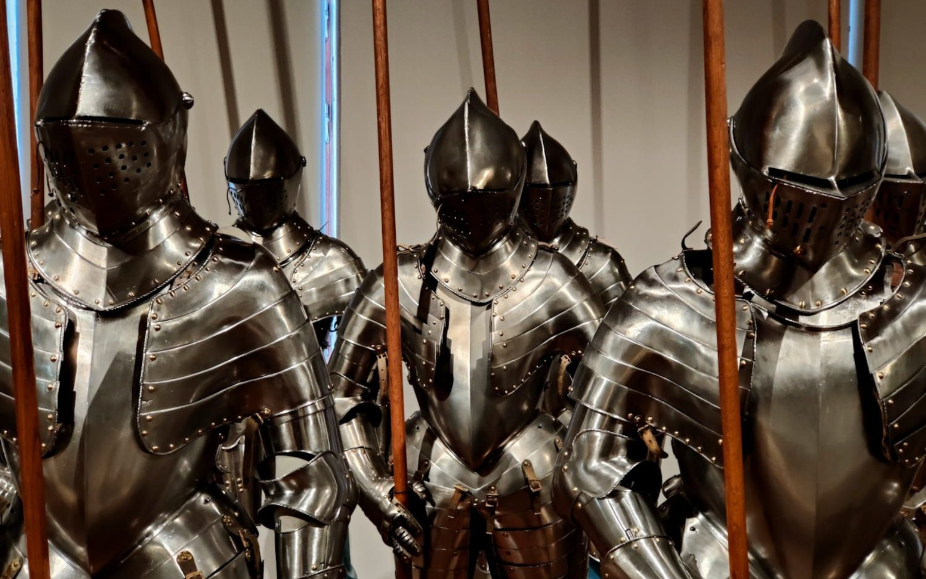 Row of shiny, medieval suits of armor, with helmets and spears, displayed indoors.