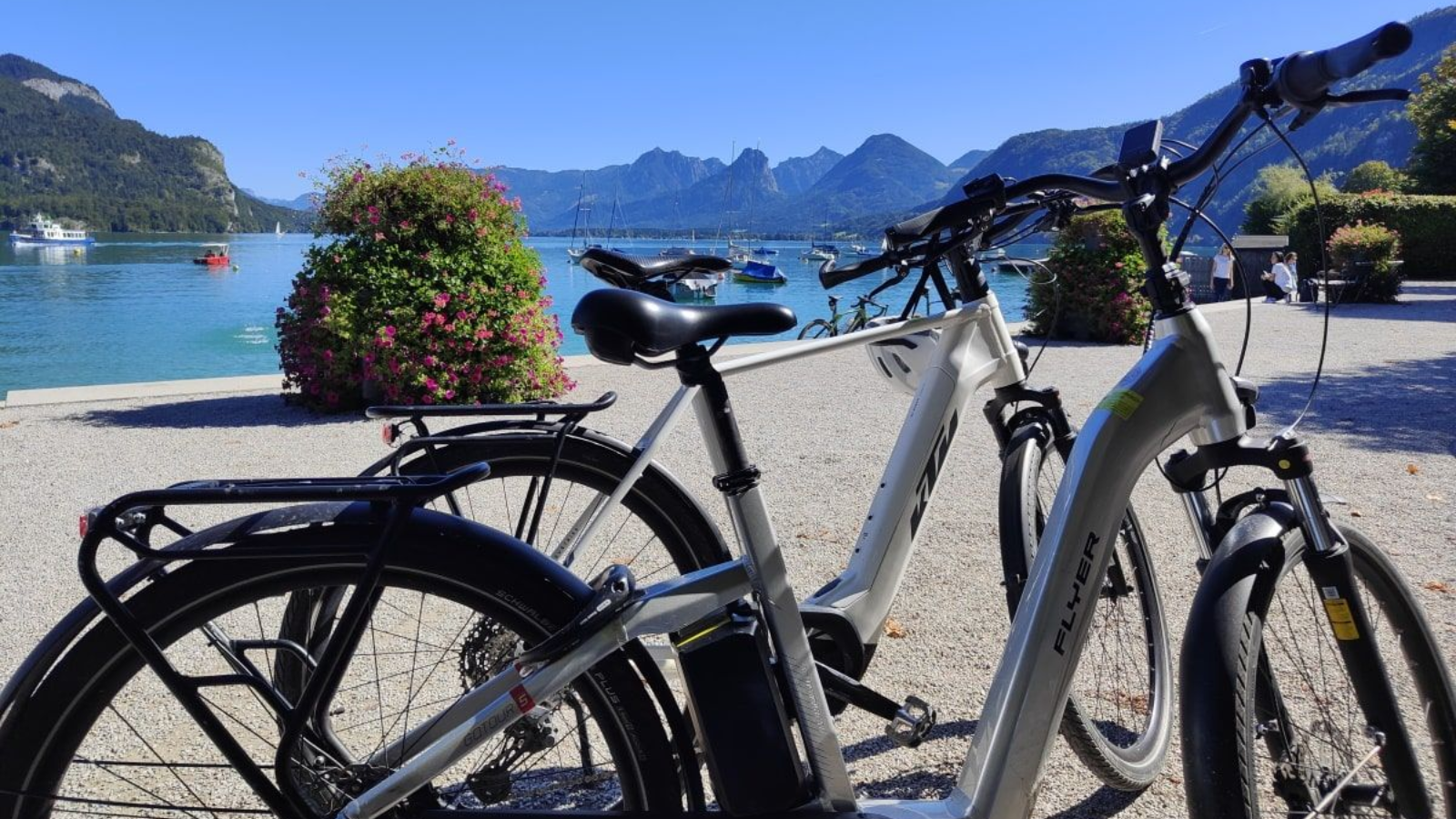 Two electric bikes parked by a lake with mountains in the background under a clear blue sky.