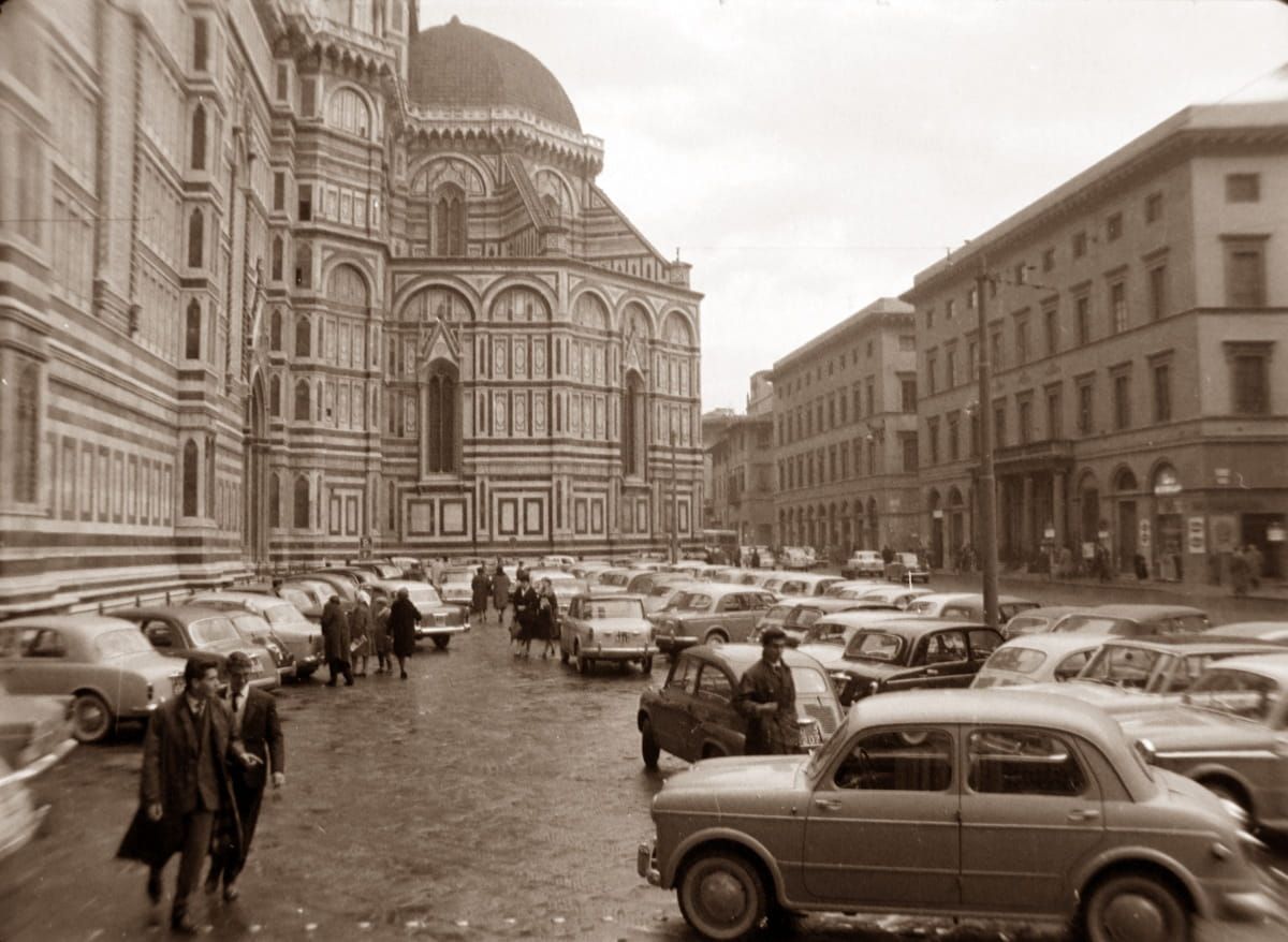 A vintage street-level view of Florence Cathedral and cars parked on the cobblestone square.