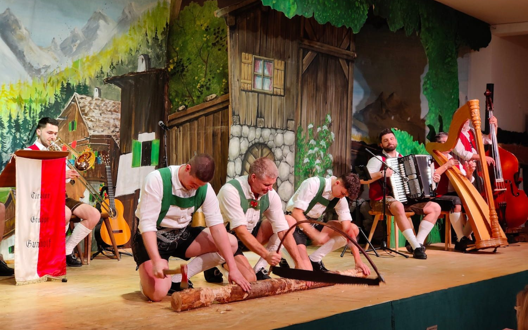 Men sawing a log on stage, traditional Bavarian setting. Musicians with accordion, harp, and bass.