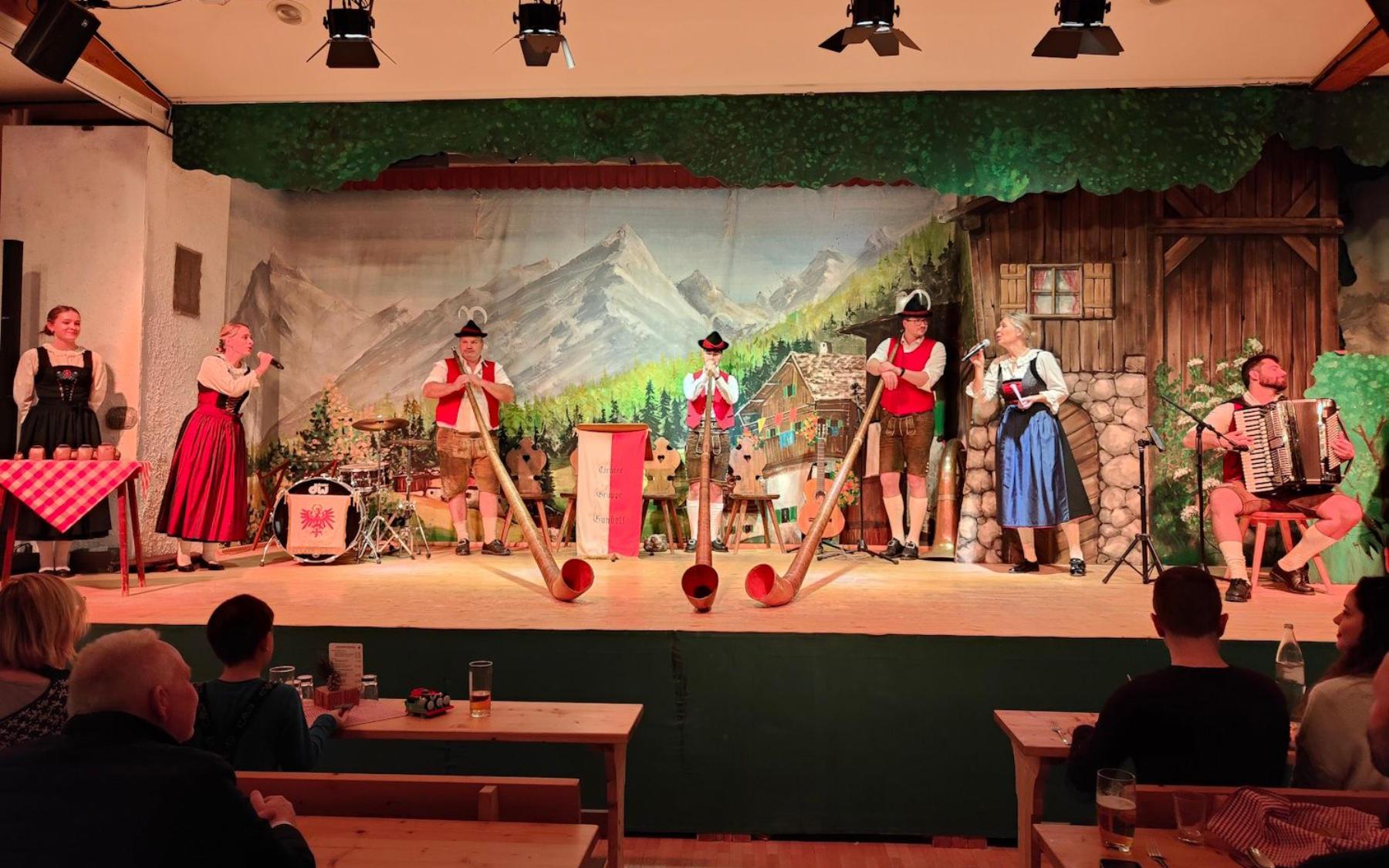 Musicians in traditional attire perform on stage, Alpine backdrop, playing instruments. Audience seated at tables.
