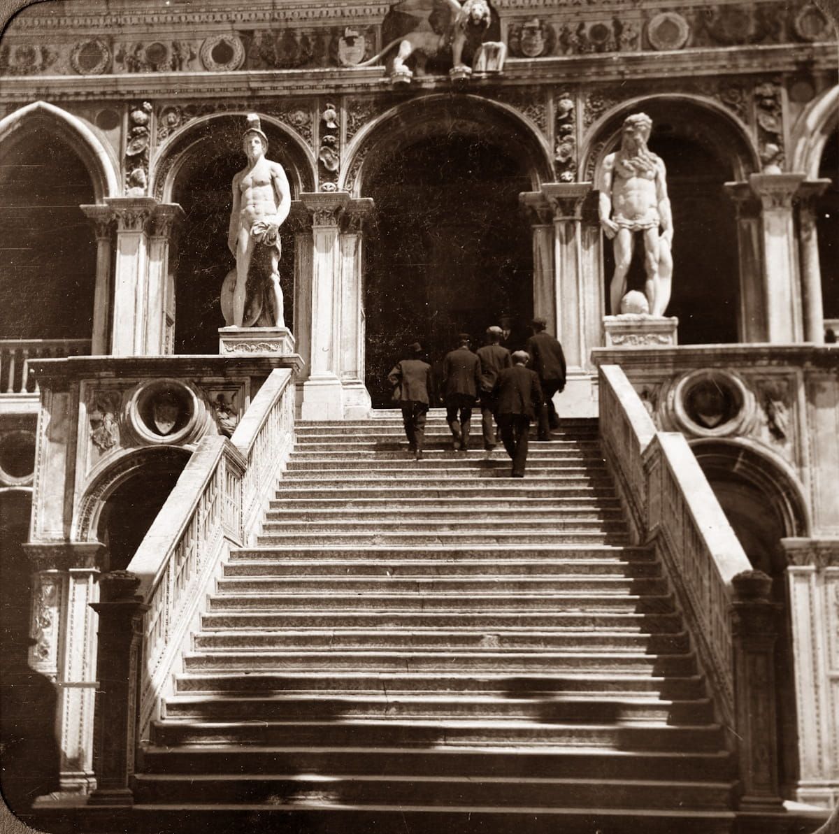People ascend the grand stone staircase, flanked by classical statues, inside the courtyard of the Doge's Palace, Venice.