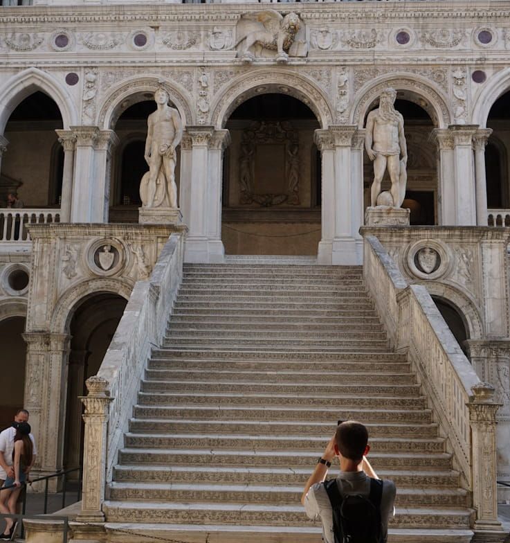 A person stands at the base of the ornate stone Giants' Staircase at the Doge's Palace in Venice, taking a photograph.