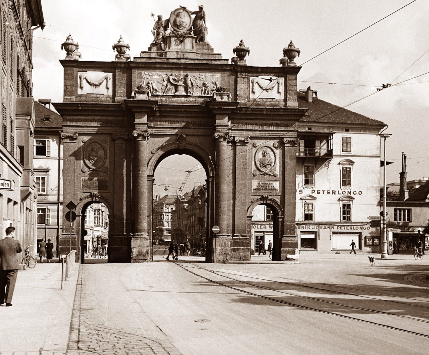 Triumphal arch with ornate details, framing a street. People walk on the road. Buildings on each side.