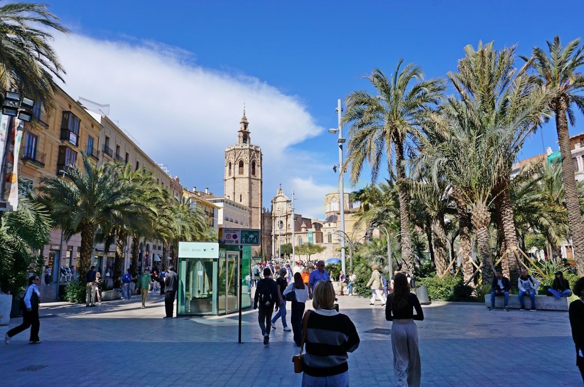 Plaza in Valencia, Spain, with cathedral tower, palm trees, and pedestrians on a sunny day.
