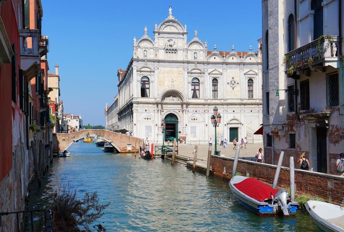 Ein Kanal in Venedig führt zu einem prächtigen, weißen Steingebäude mit Bogenfenstern unter einem strahlend blauen Himmel.