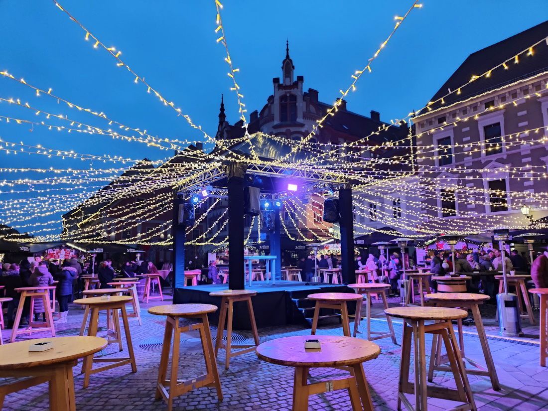 Outdoor stage with string lights, tables, and people in a European town square at dusk.