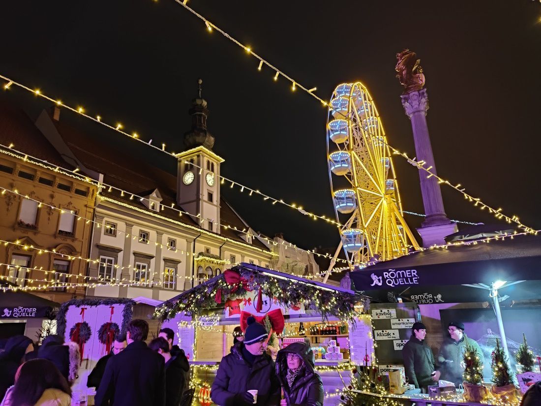 Night view of a Christmas market with a Ferris wheel, stalls, and buildings lit up. People are present.