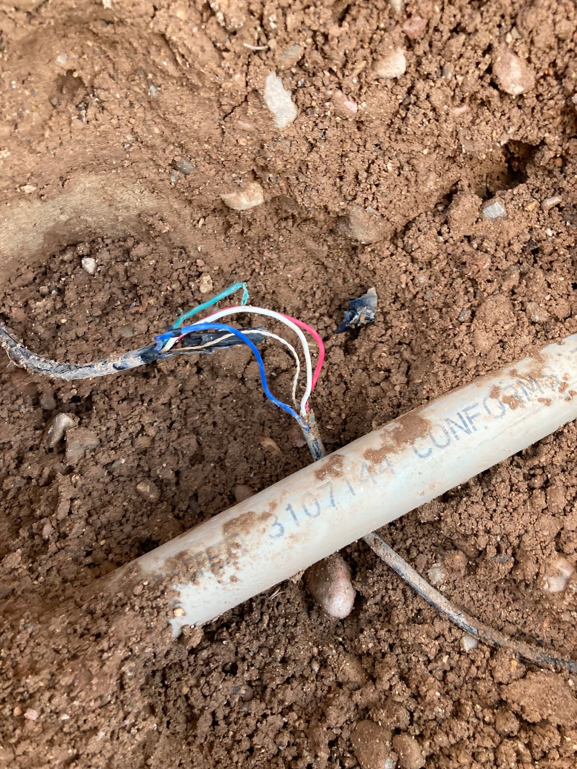 Exposed wires and irrigation pipe in dirt. White, blue, red, and green wires emerge from the ground.