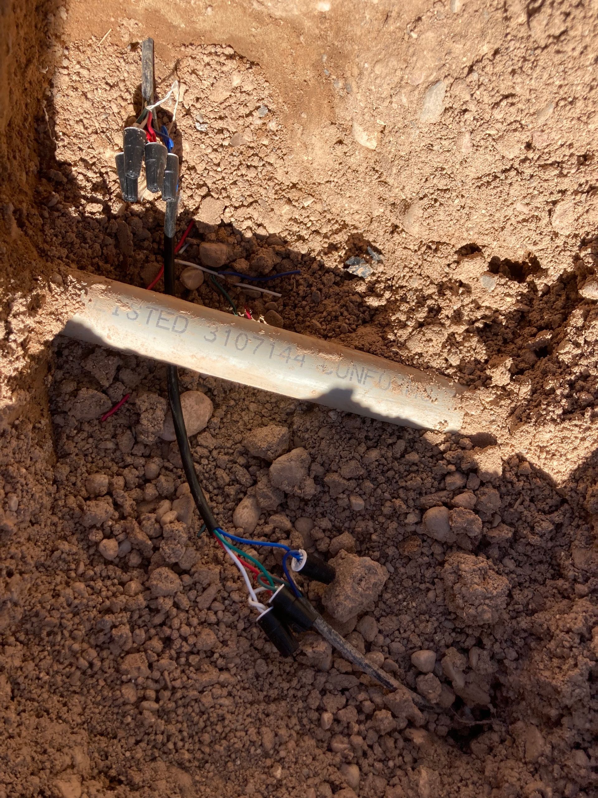 Wires and a white pipe in a dirt trench. Several colored wires exposed and bundled.