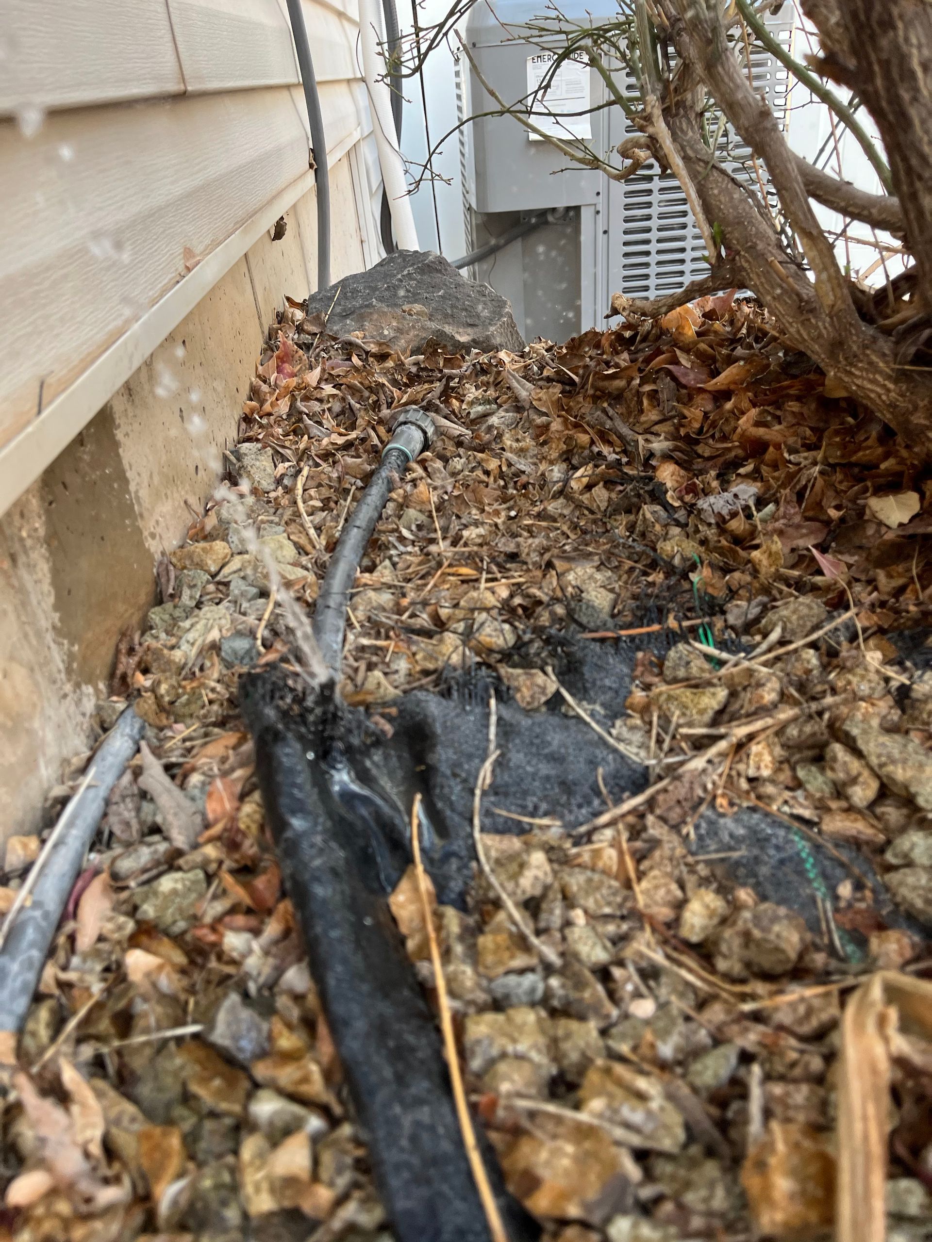 Gravel and debris near a building foundation, with black pipes and a small tree.