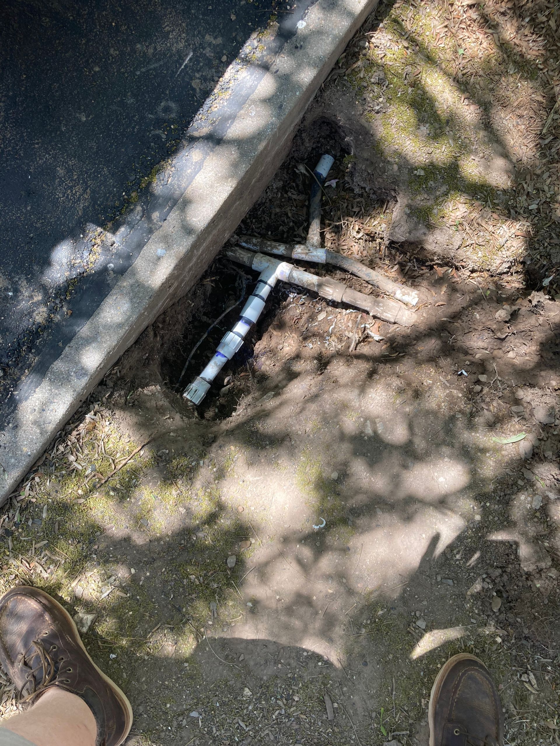 Black and white PVC pipes extending from the ground next to a dark paved surface, with grass and shadow, seen from above.