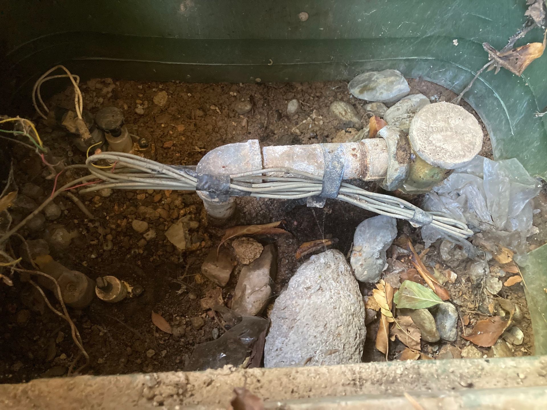 Green box containing a rusted metal pipe, wires, and small rocks in soil.