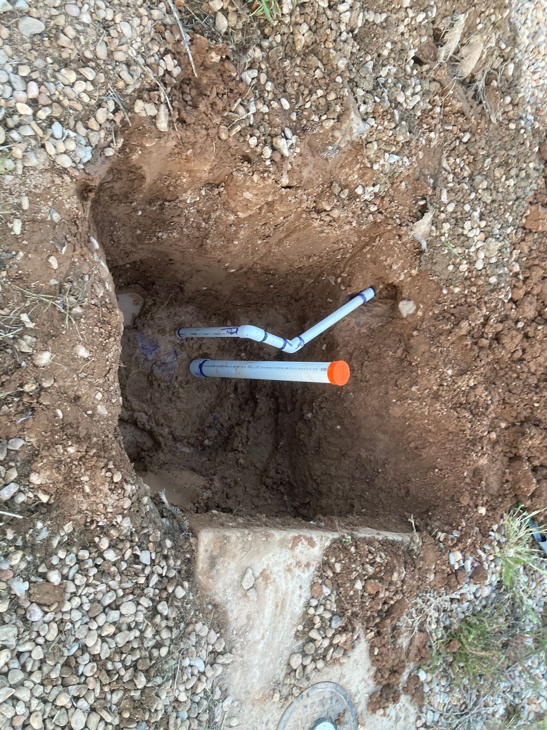A small square pit in the ground reveals two water pipes, one with an orange cap, in a gravelly setting.