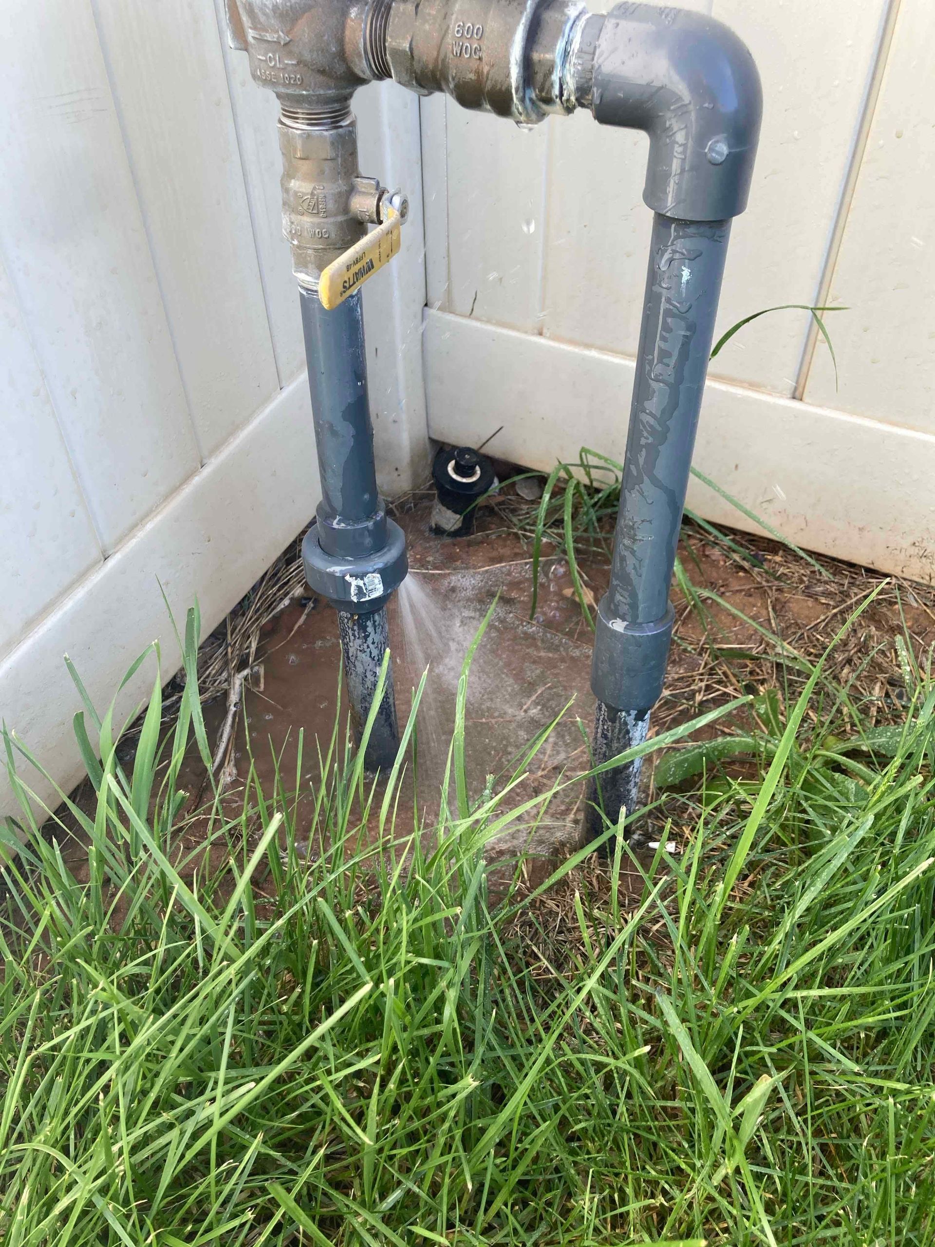 Water spraying from a damaged pipe near a white fence, on grass, in an outdoor setting.