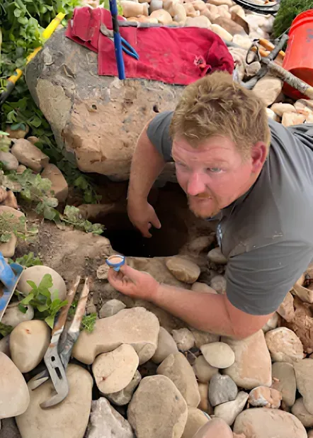 Man reaching into a dark hole surrounded by rocks, holding a small container.