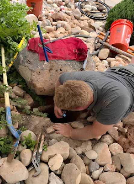 Man leans into a hole in a rock garden with tools and stones surrounding him.