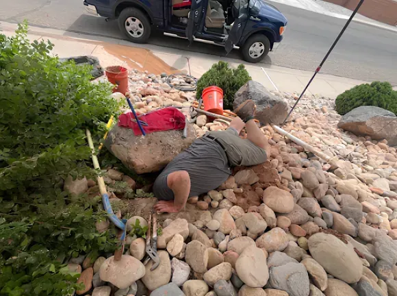 Man working in rocky garden bed, head and arms in the ground, blue truck parked nearby.