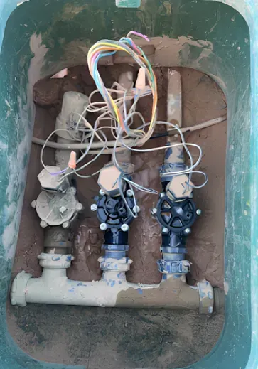 Irrigation valves with wiring inside a green plastic box, covered in mud.