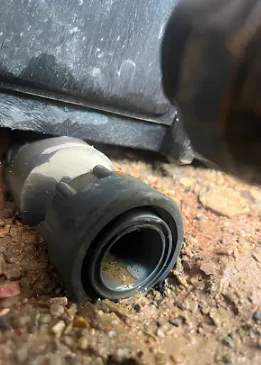 Close-up of a gray plastic pipe fitting on reddish-brown dirt beneath a dark structure.