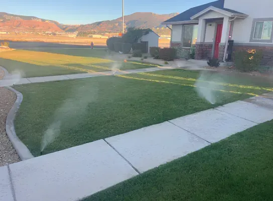 Sprinklers watering a green lawn in front of a house with a brick walkway.