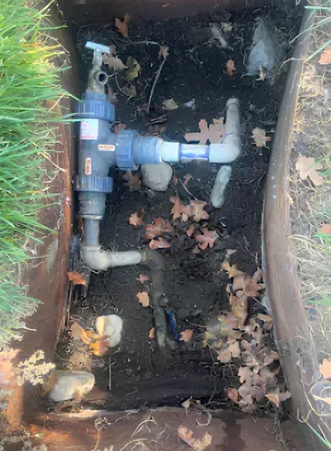 Water valve system in a dirt-filled box, connected to pipes and a spigot, surrounded by leaves and grass.