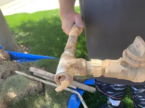 Person holding a broken, mud-covered irrigation pipe outdoors, near grass and landscaping materials.