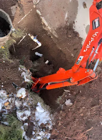 A small orange excavator digging in brown soil near a white pipe and a concrete structure.