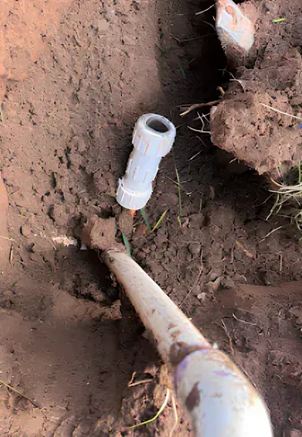 A white PVC pipe fitting and shovel in a dirt trench.