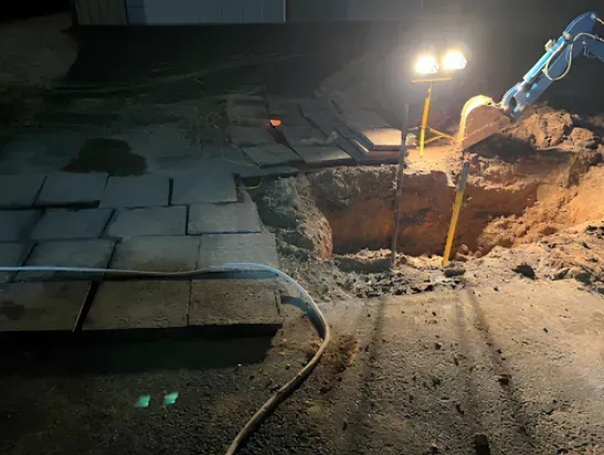 Excavation work at night, next to pavers and building. Bright work light illuminates dirt, machinery.