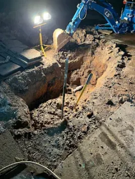 Excavation site at night with digger, spotlights, and tools in a deep hole in asphalt.