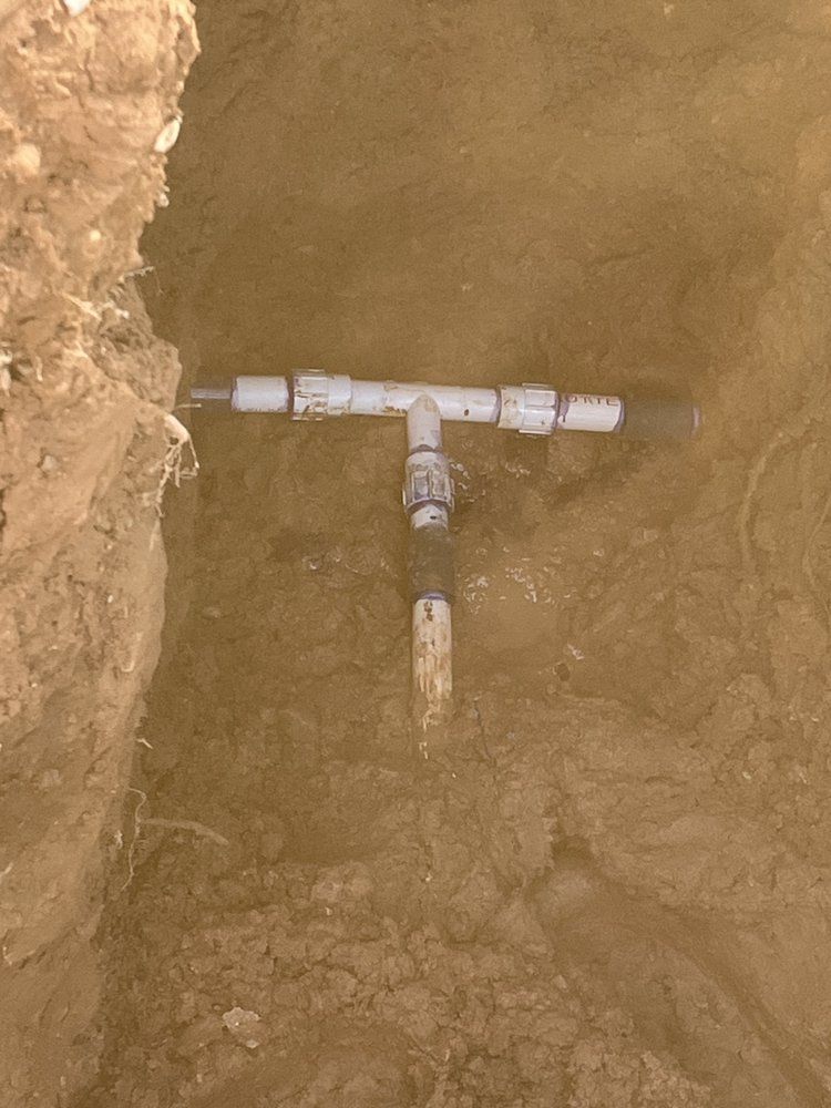White PVC pipes connected in a trench. A T-shaped fitting is connected to two straight sections, buried in dirt.
