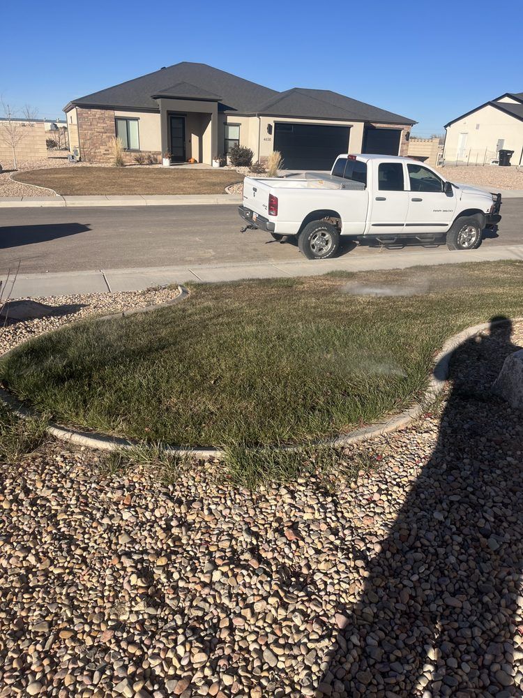 White truck driving on a street in front of a house with a lawn being watered; sunny day.